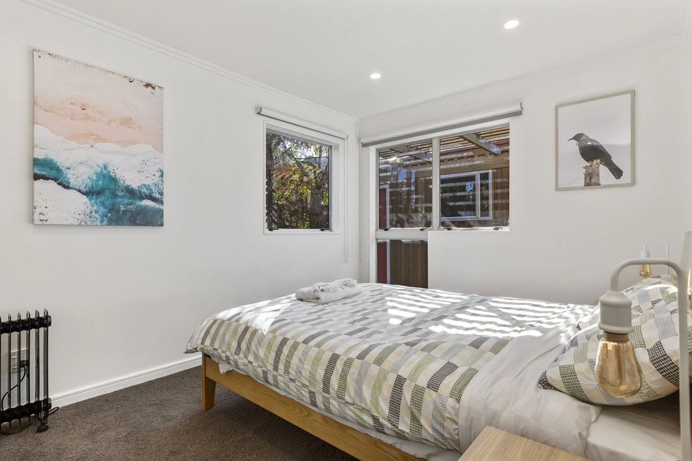 A bedroom with a bed, two windows, and artwork on the walls, sunlight casting shadows on the bed.