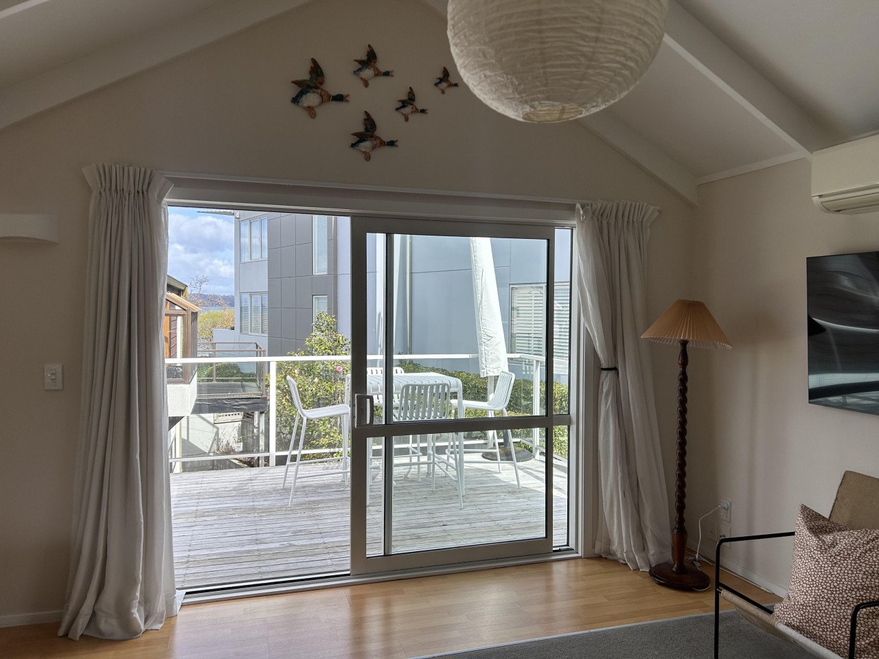 Living room with sliding glass door leading to a balcony, outdoor table with chairs, air conditioning unit, floor lamp, flat-screen TV, and a ceiling paper lantern.