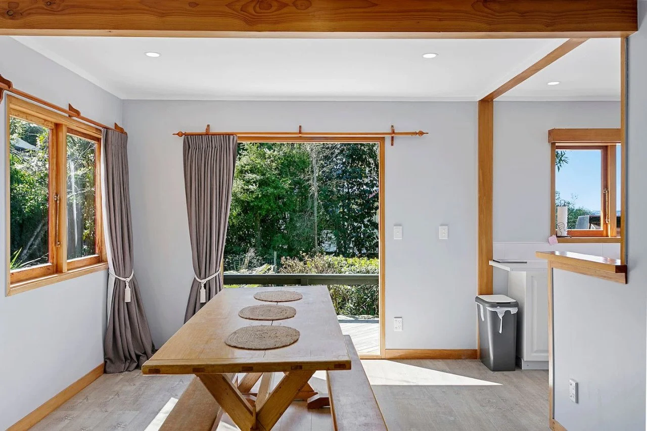 Bright dining area with a wooden table, gray curtains, and large windows showing greenery outside.