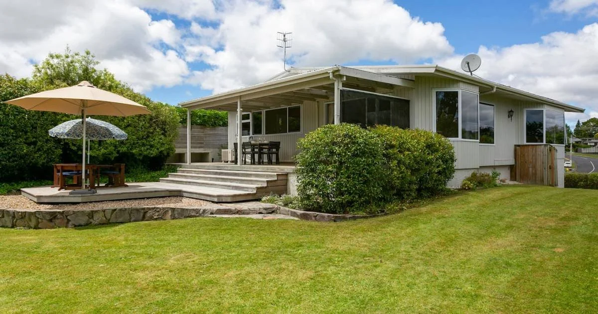 A white house with a large front porch, surrounded by green bushes and grass, with a patio area featuring an umbrella and outdoor furniture, under a partly cloudy sky.