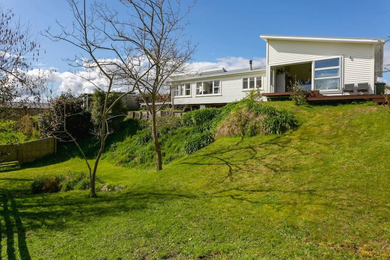 A white house on a grassy hill with a deck, leafless trees, and a blue sky with clouds.