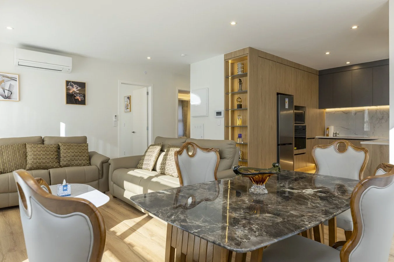 Modern open-plan living room and kitchen with beige sofas, a dining table with a marble top, and wood accents.