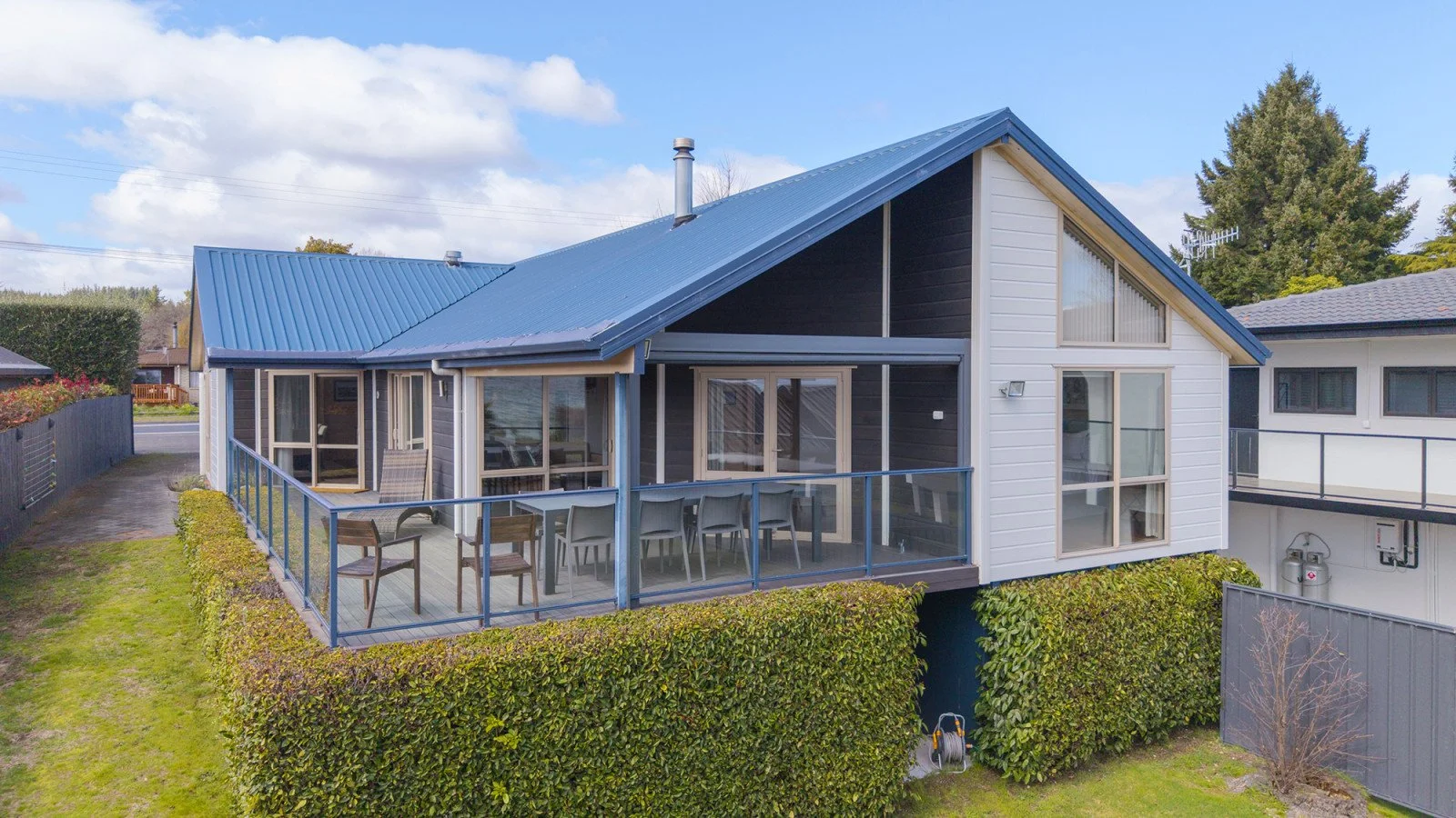 Modern house with a blue metal roof, large windows, and a balcony with outdoor furniture, surrounded by greenery.