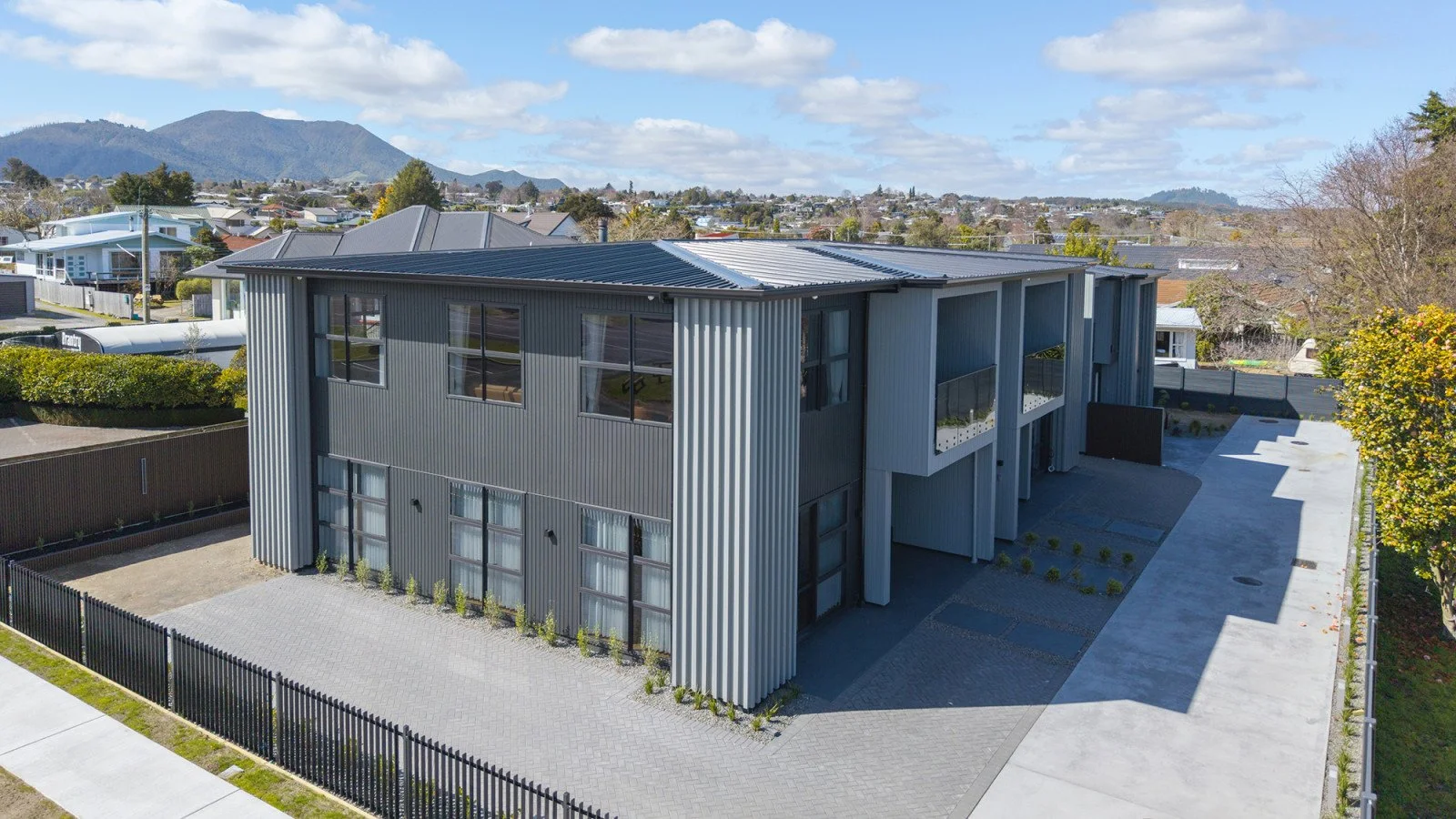 A modern two-story residential building with gray metal siding in a suburban neighborhood, surrounded by a black metal fence, with a paved driveway and small landscaped areas. Mountains and other houses are visible in the background.