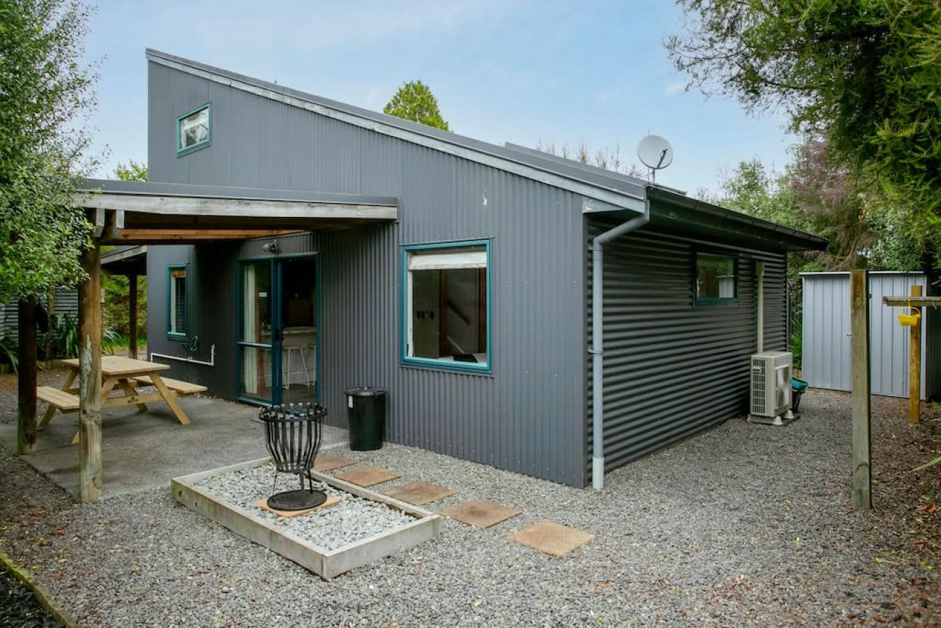 A modern house with a grey metal exterior, a sloped roof, and several windows, surrounded by a gravel yard with outdoor furniture, a fire pit, and trees.