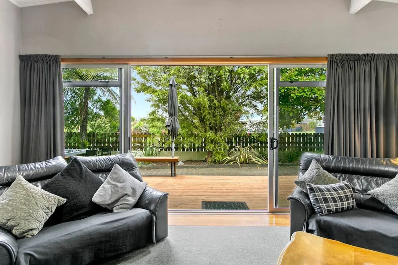Living room with two black leather sofas and gray cushions, large sliding glass door opening to a wooden deck outside with trees and a garden.