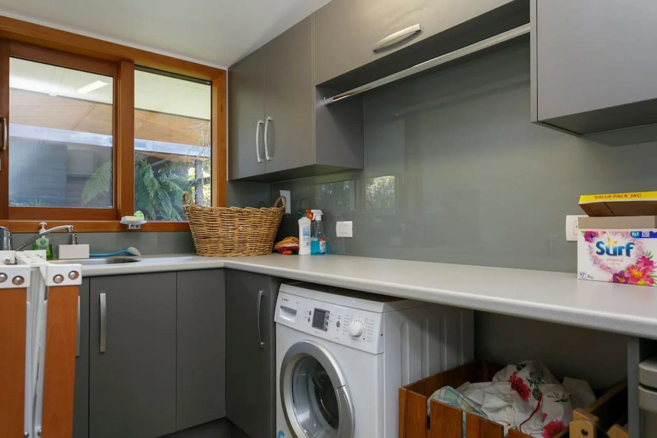 Laundry room with grey cabinets, a washing machine, a window with a view of greenery, a basket, and some cleaning supplies.