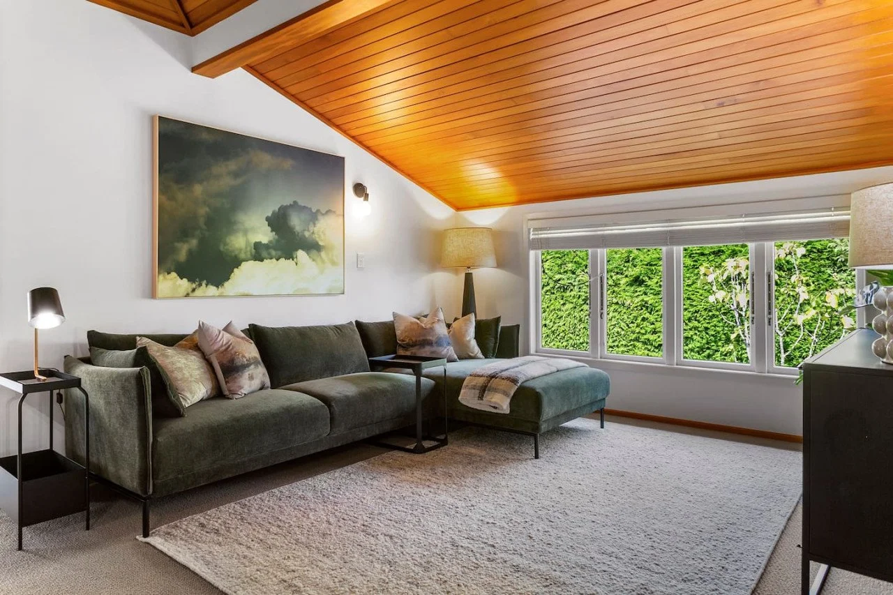 Living room with a dark green sofa, pillows, a side table with a lamp, a large window with greenery outside, and a wooden ceiling.