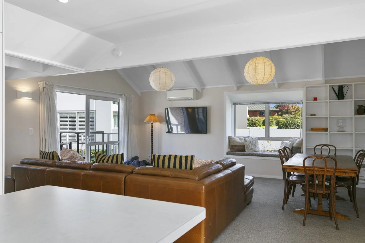 Living room with a leather sofa, a wall-mounted TV, a window seat with cushions, a dining table with chairs, and a large window showing outdoor views.