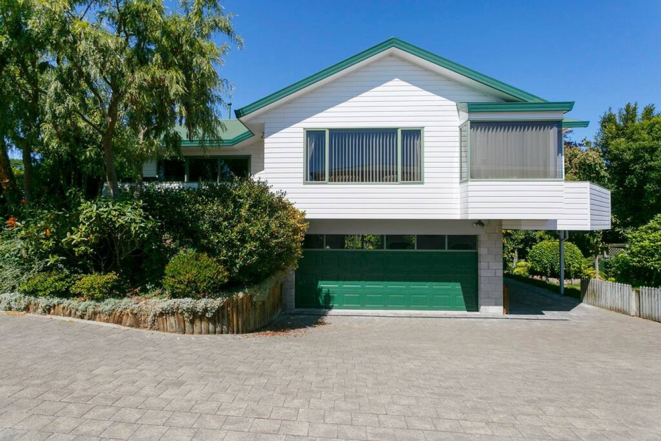 A two-story house with white siding, green garage door, and green roof. Surrounded by greenery and a paved driveway.