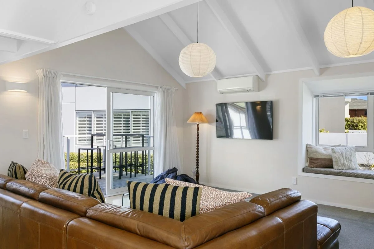 Bright living room with a brown leather sectional sofa, striped and patterned pillows, a floor lamp, a wall-mounted TV, and large windows with white curtains.