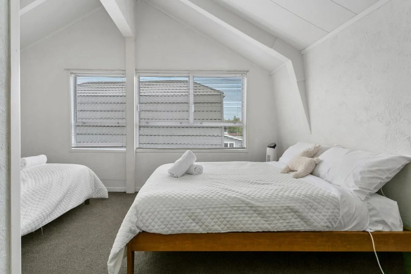 A bedroom with white walls, a large bed with white bedding and pillows, a rolled towel on the bed, and a window with blinds showing a neighboring house and blue sky.