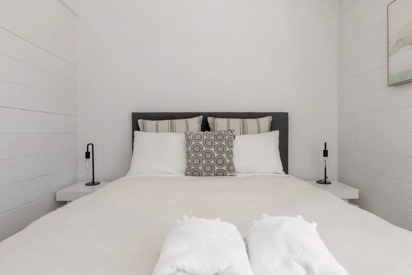 Clean and modern bedroom with white walls, a gray upholstered headboard, white bedding, two bedside lamps, and decorative pillows.