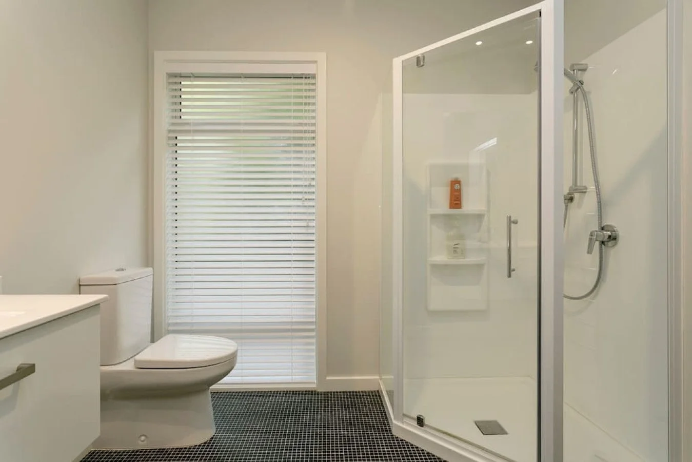 Modern bathroom with a white toilet, a shower with glass doors, and a window with white blinds.