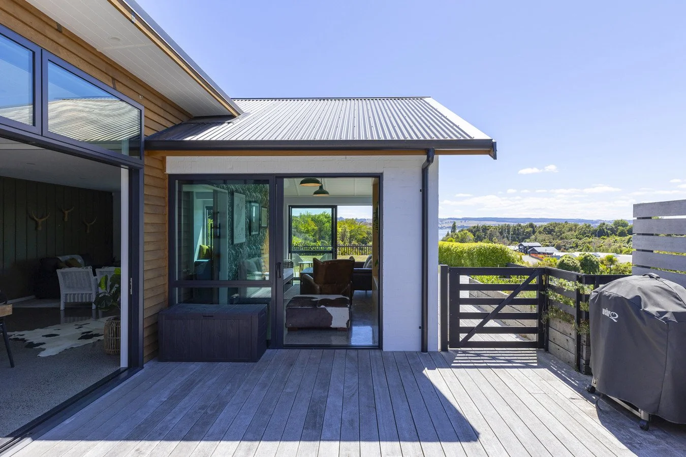 View of a wooden balcony with a sliding glass door leading to a living room, overlooking a scenic landscape with trees and a river in the distance. There is a BBQ grill covered on the right and outdoor seating visible inside.