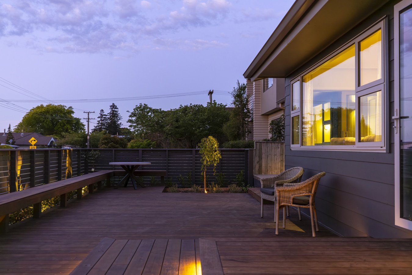 A wooden deck with outdoor furniture including a round table and two wicker chairs, illuminated by soft evening lighting with trees and houses visible in the background, and a house window glowing with warm light.