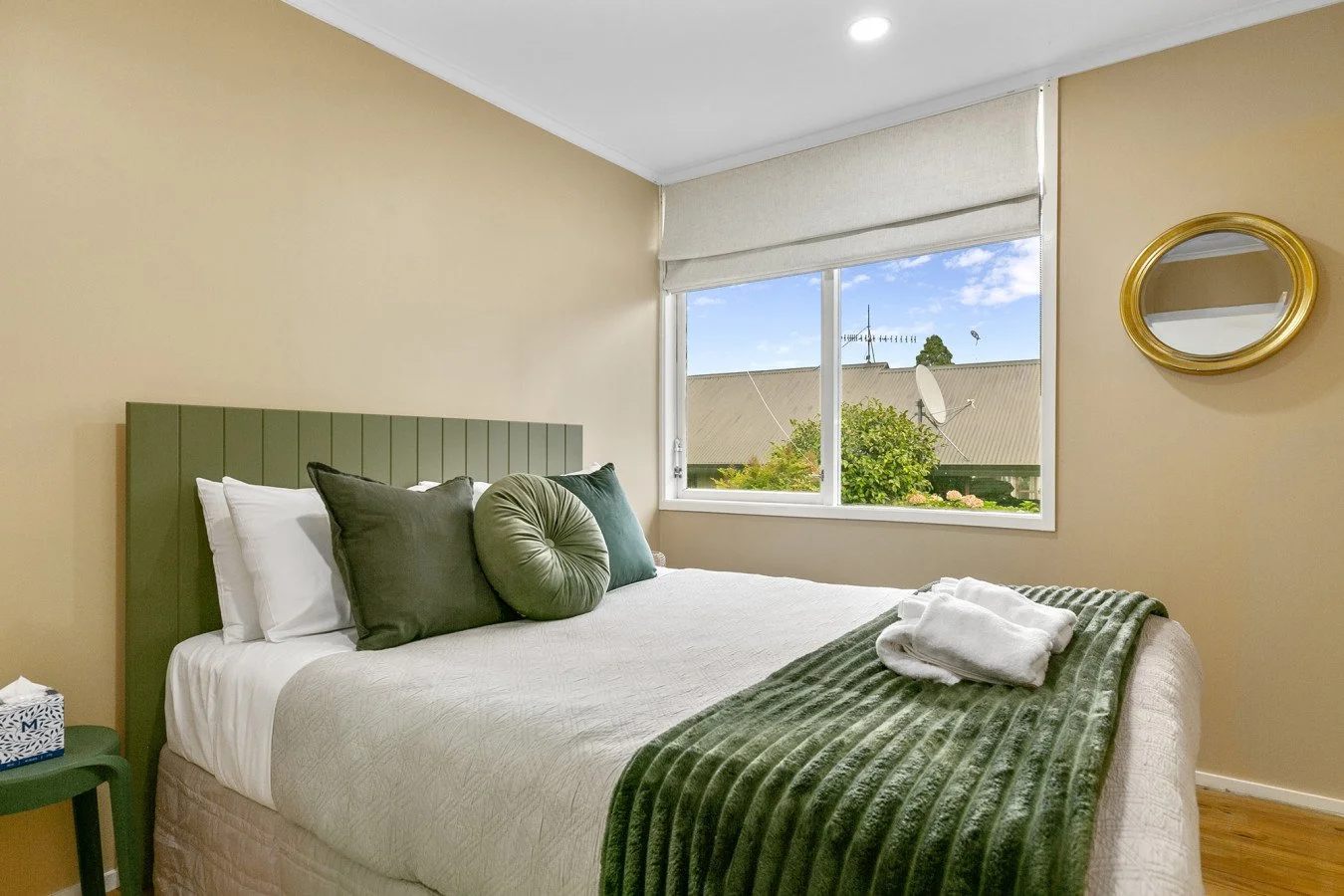 Bedroom with a green headboard and pillows, window with outside view, round mirror on wall, and neatly folded towels on bed