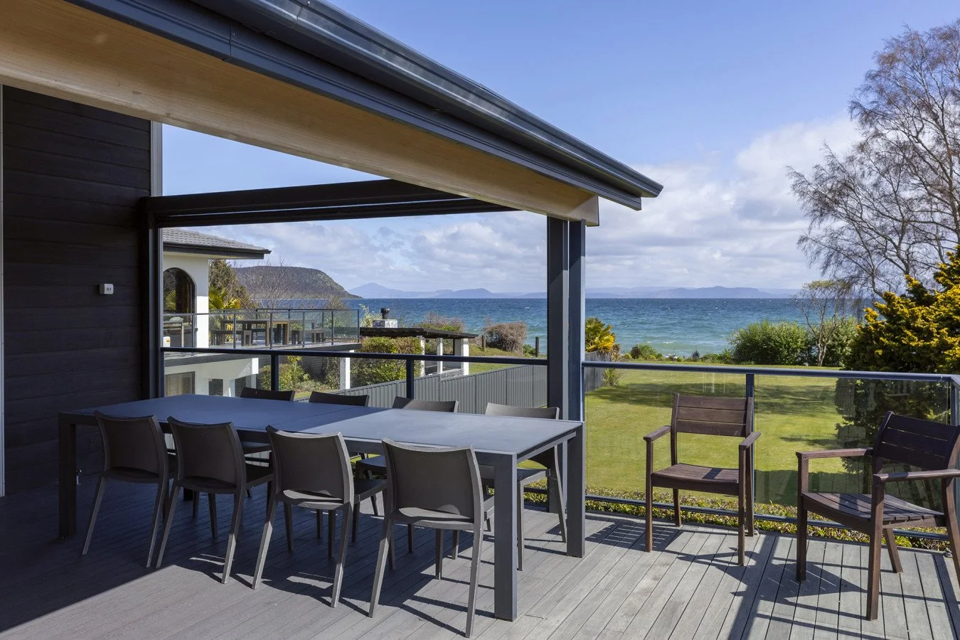 Balcony with outdoor dining table and chairs overlooking grassy yard and ocean in the distance under partly cloudy sky.
