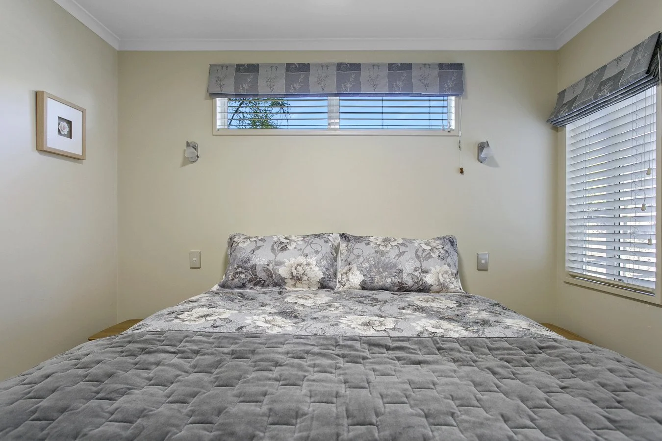A neatly made bed with floral-patterned pillows and a gray quilt in a bedroom with cream-colored walls, a framed picture on the left wall, two small wall lamps, and windows with blinds and valances.