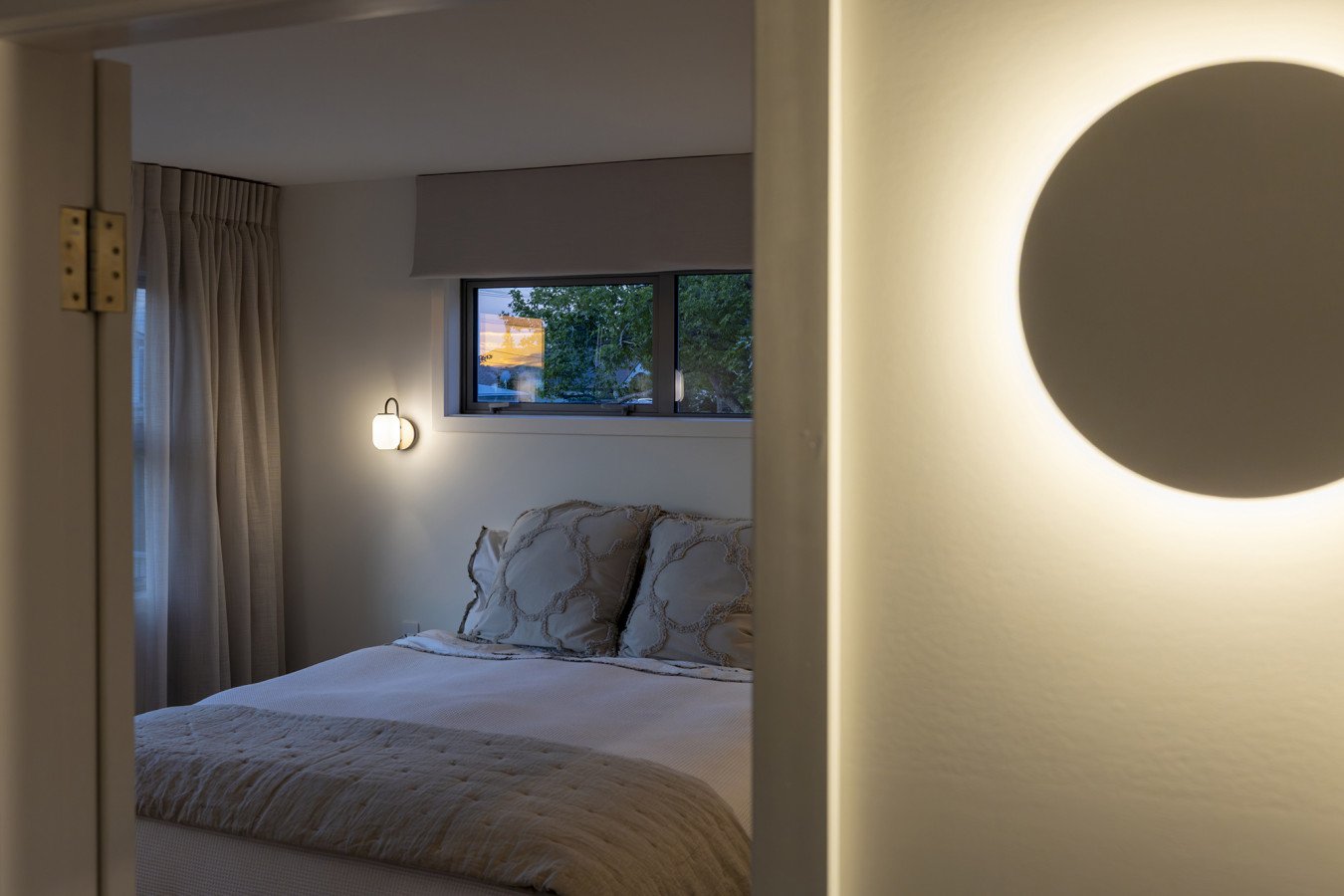A cozy bedroom with a bed, decorative pillows, a window showing trees outside, and wall-mounted lights