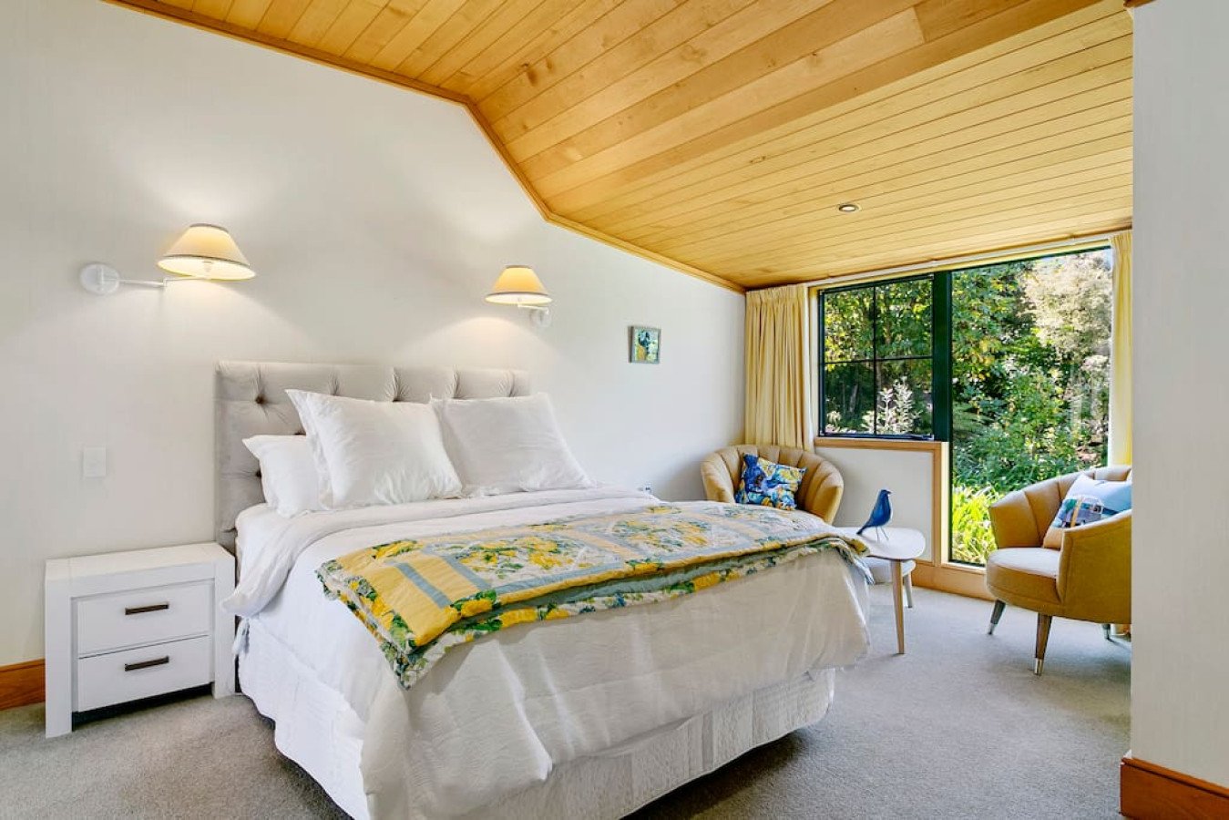 Bright bedroom with a large bed, white bedding, and multiple pillows, near a window with yellow curtains. Two yellow chairs with cushions, a small white side table with a bird figurine, and wall-mounted lamps.