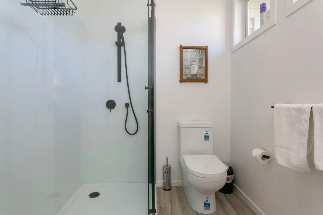 A bathroom with a walk-in shower and a toilet. The shower has a black handheld showerhead and a glass partition. The toilet has a white tank and a closed lid. There is a towel hanging on a rod and a small waste bin. The wall has a framed display with
