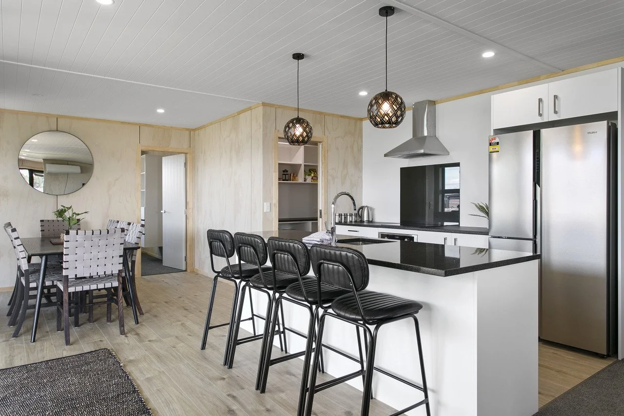Modern kitchen with white cabinets, stainless steel refrigerator, black countertop island with four black barstools, stainless sink, and a stainless range hood. Adjacent dining area with a table and six woven pattern chairs, a round mirror on the wal