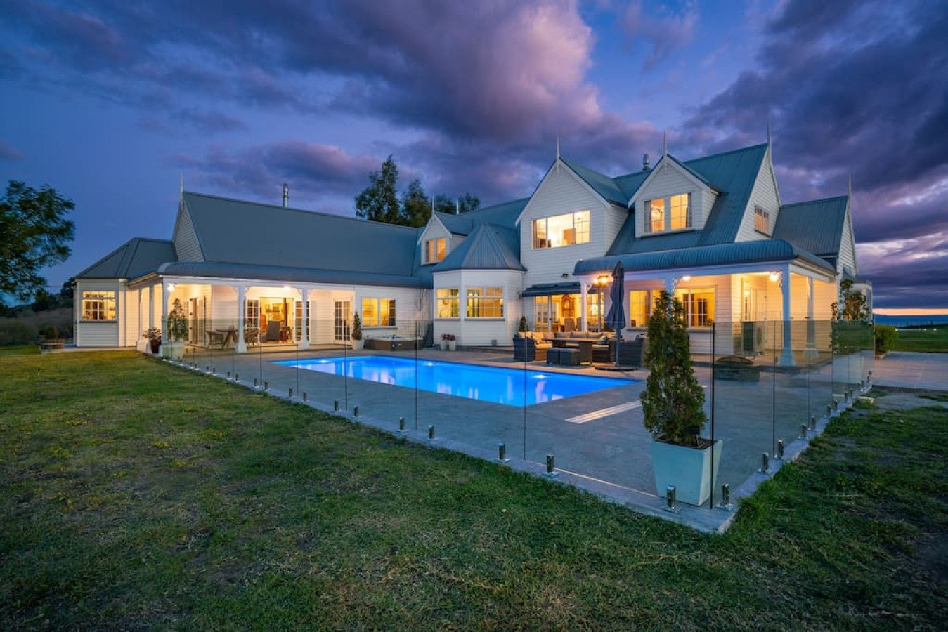A large luxurious house at dusk with lights on, a lit swimming pool, and a fenced backyard with patio furniture.