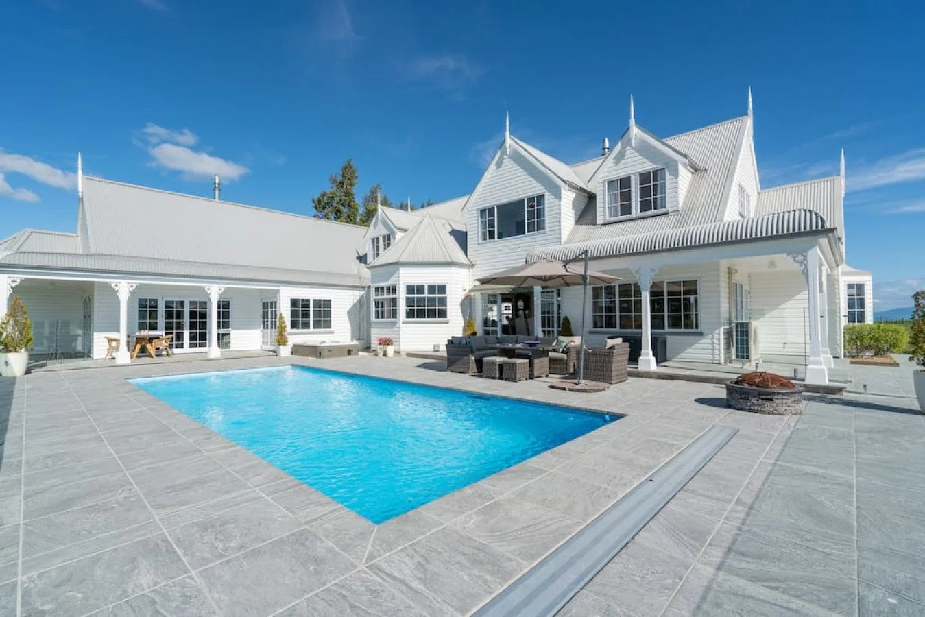 Large white house with steep roof, multiple windows, and a backyard pool under a blue sky.