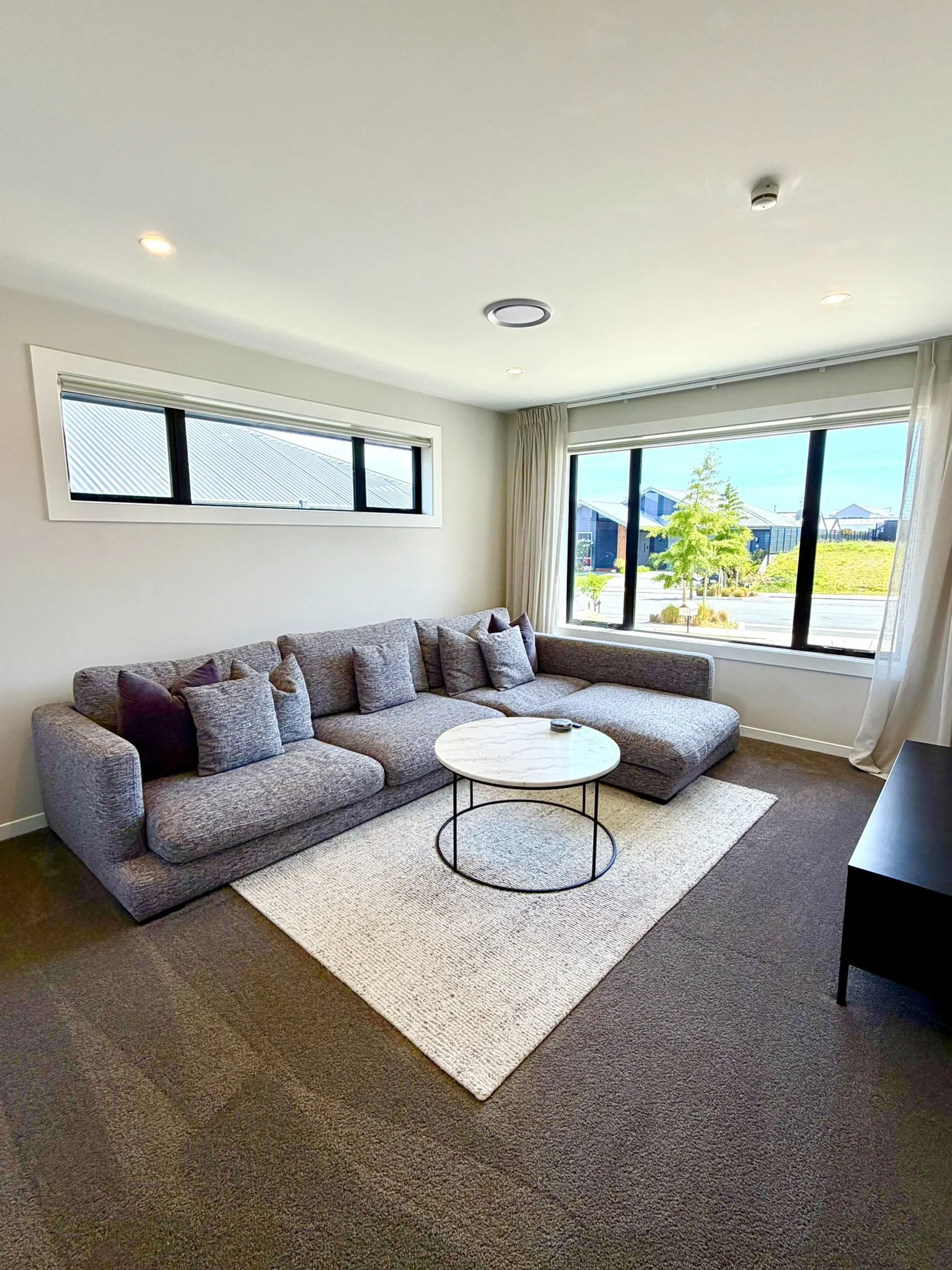 Living room with a large gray sectional sofa, marble-top round coffee table, beige area rug, large windows with curtains, and ceiling lights.