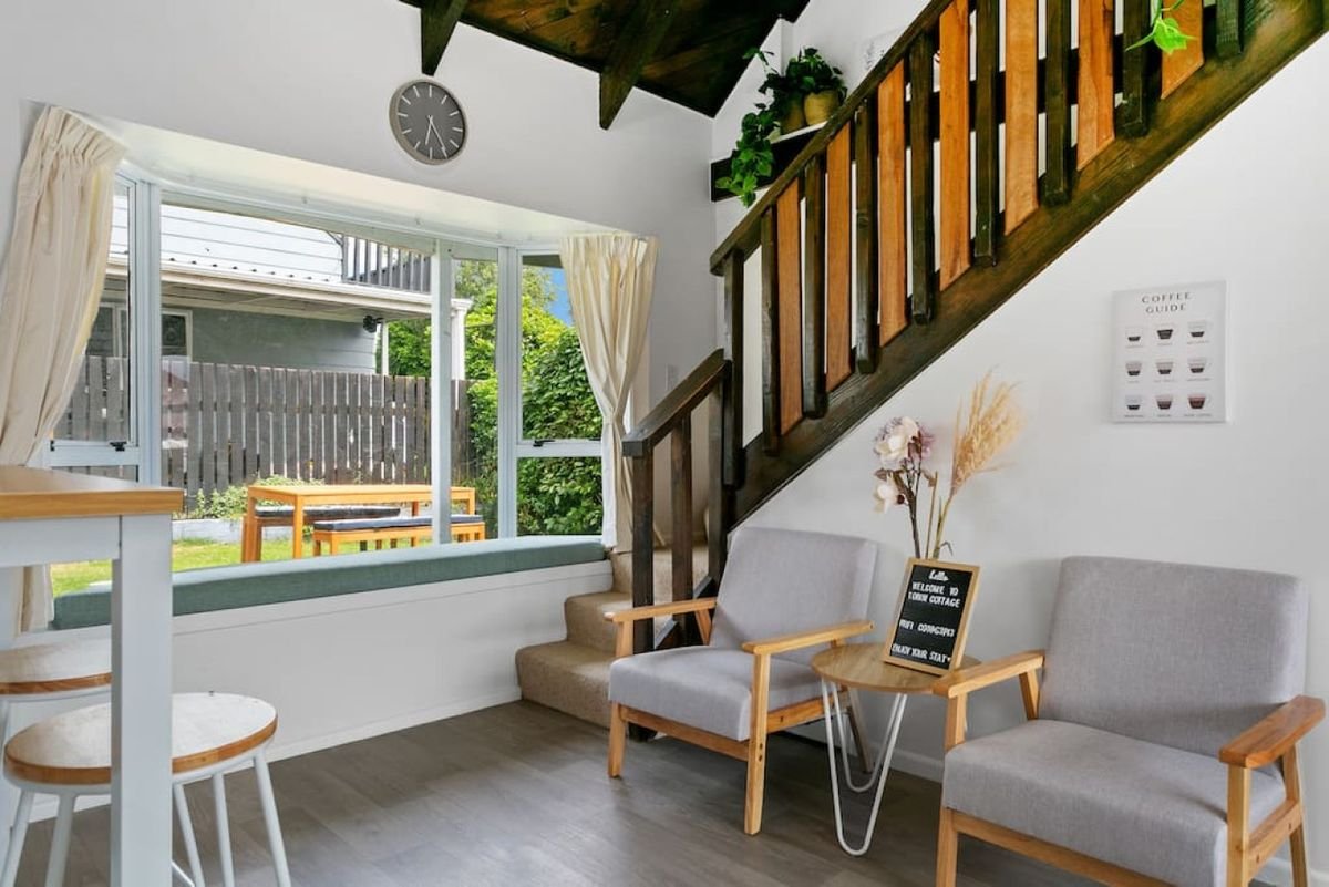 Living room with a large window seat, two wooden armchairs with light gray cushions, a small round wooden table with a blackboard sign, potted plants on a staircase railing, and a wall with a coffee guide poster.