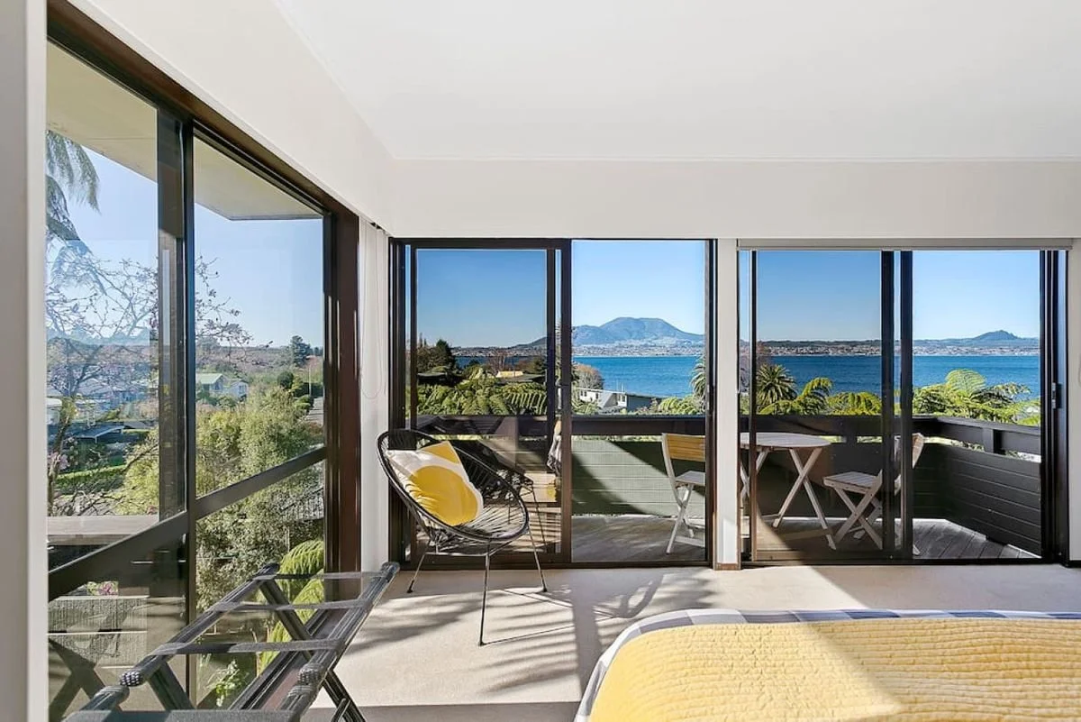 View from a living room with large glass sliding doors opening to a balcony overlooking the ocean, with mountains in the background, and interior furniture including chairs, a bed, and a yellow blanket.
