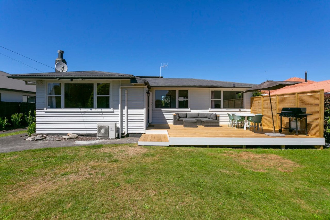 Backyard with a wooden deck, outdoor furniture, umbrella, barbecue grill, and green lawn under a clear blue sky.