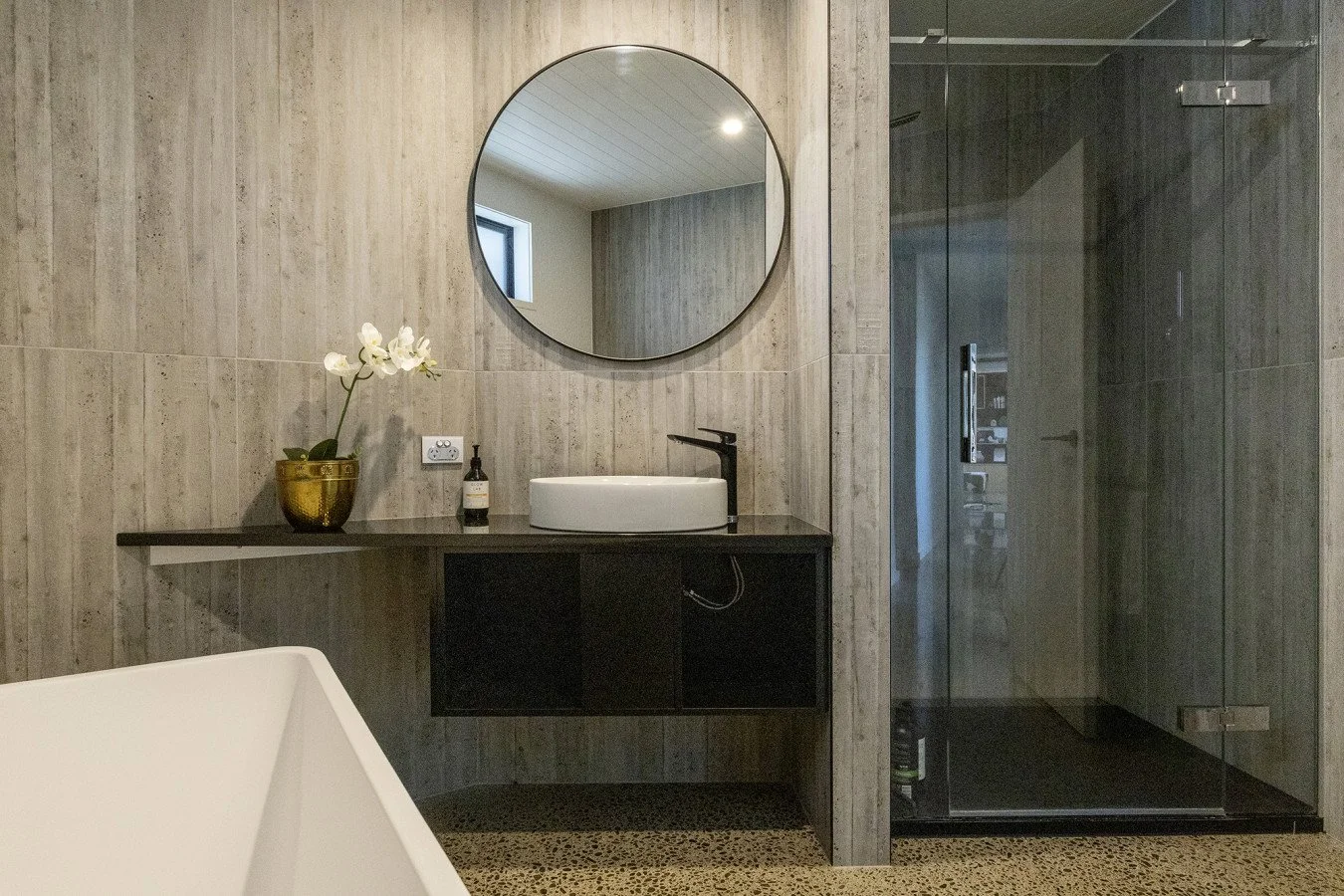 Modern bathroom with a round mirror above a vessel sink, black countertop, and a black floating cabinet, with a shower area enclosed in glass on the right.