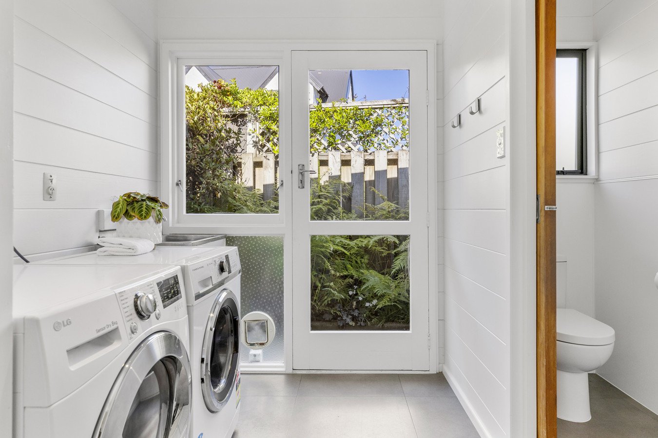 Laundry room with washing machine, dryer, and a door leading to a small outdoor area with greenery.