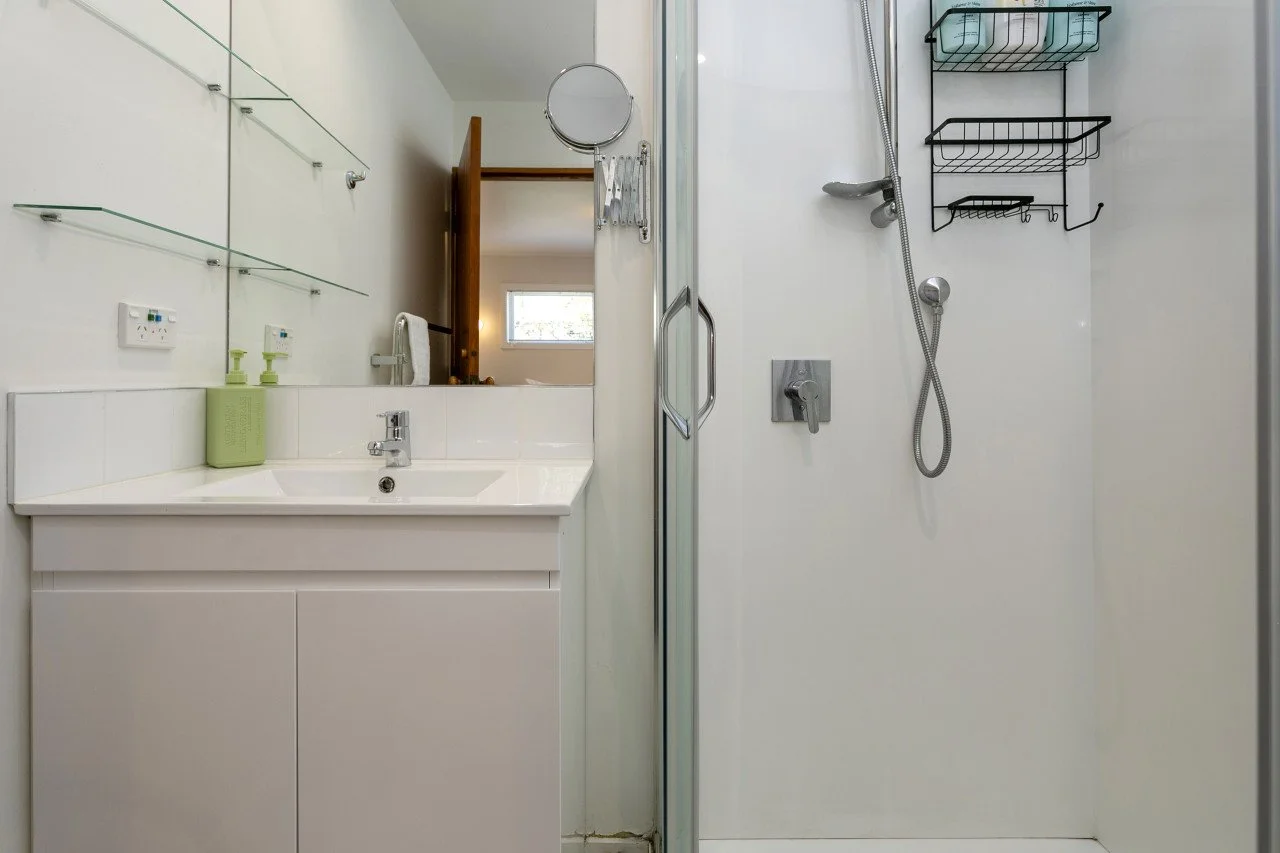 A modern bathroom with a white vanity and sink, mounted mirror, and two green soap dispensers. To the right, there is a separate shower area with a glass door, a handheld showerhead, and black wire shelves for toiletries.