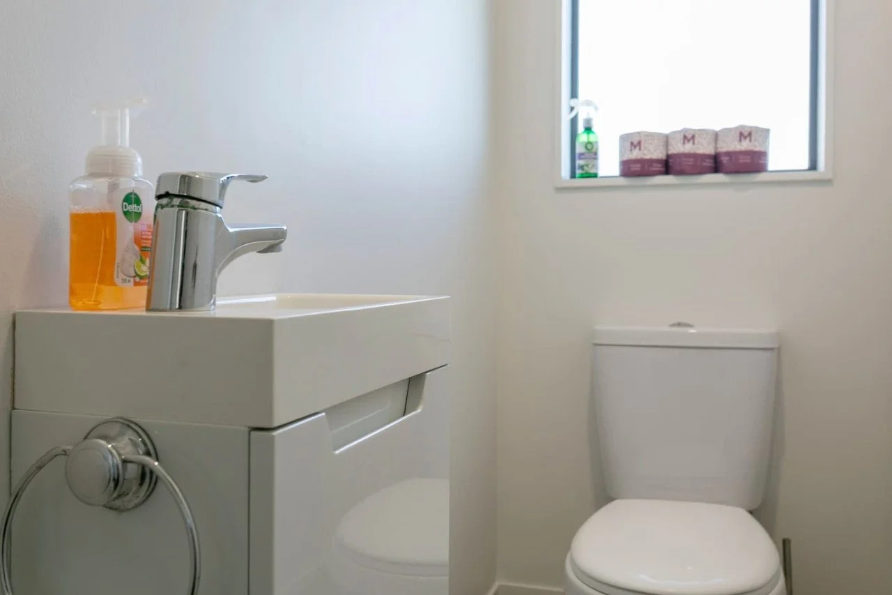 A small bathroom with a white sink, soap dispenser, and a toilet with a window above it, holding a spray bottle and tissue boxes.