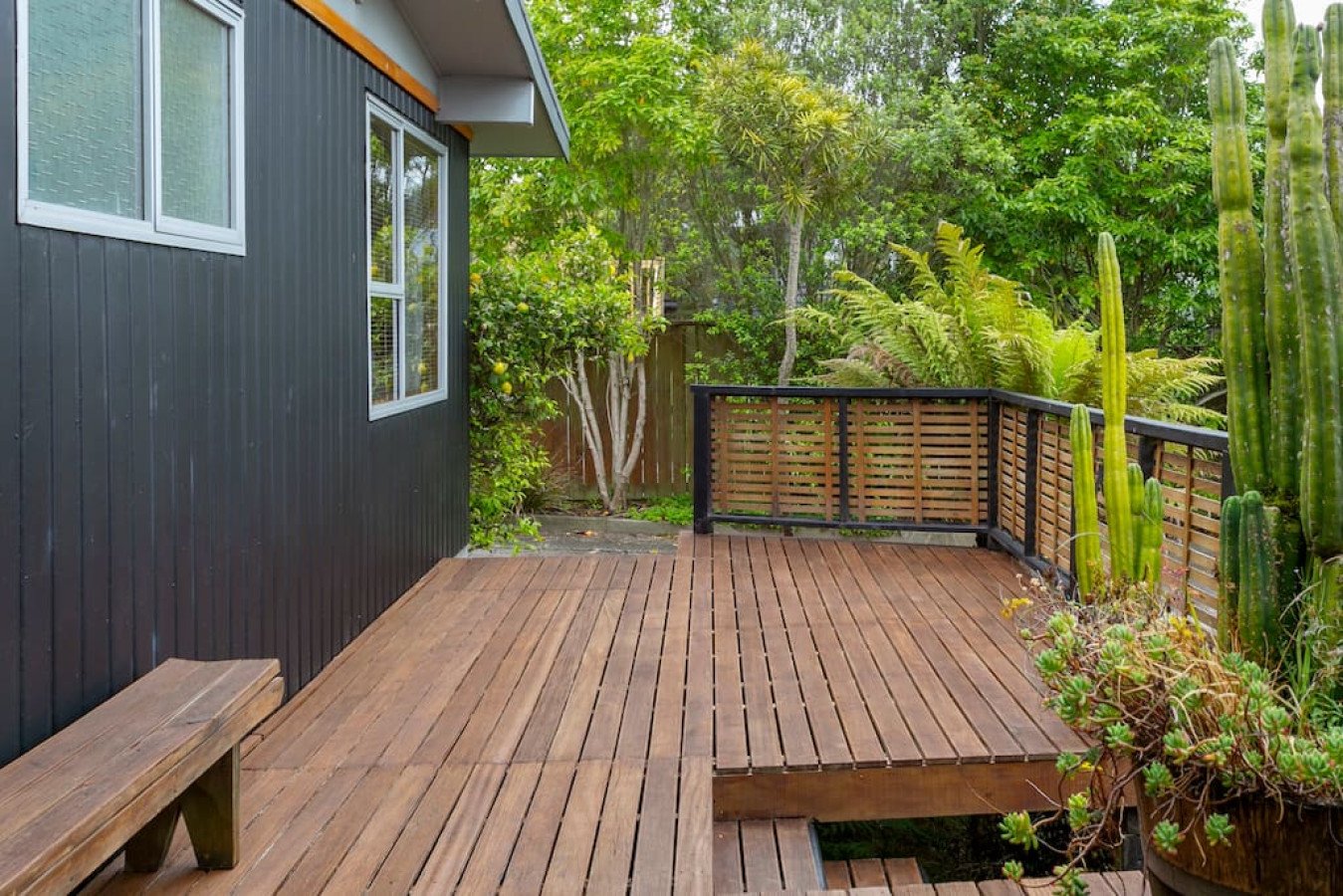 Wooden deck with black railing surrounded by lush green plants and trees, part of a modern house with black exterior walls and white-framed windows.
