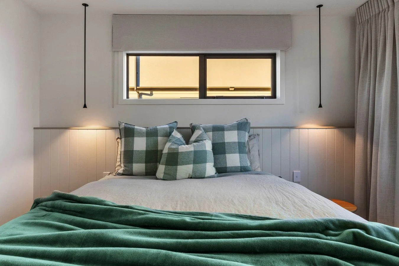 A neatly made bed with three checkered pillows, a green blanket at the foot, a window above with beige blinds, and modern lighting fixtures on each side of the bed in a bedroom setting.
