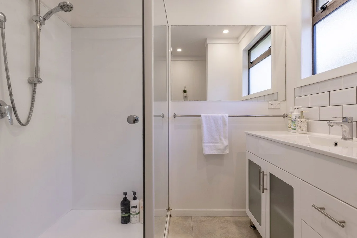 Modern bathroom with walk-in shower, white vanity, large mirror, window, and neutral-toned tiled floor.