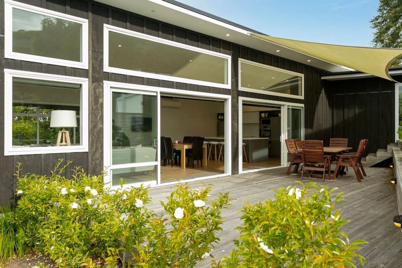 Modern house with large glass sliding doors, outdoor patio with wooden table and chairs, and lush green bushes.