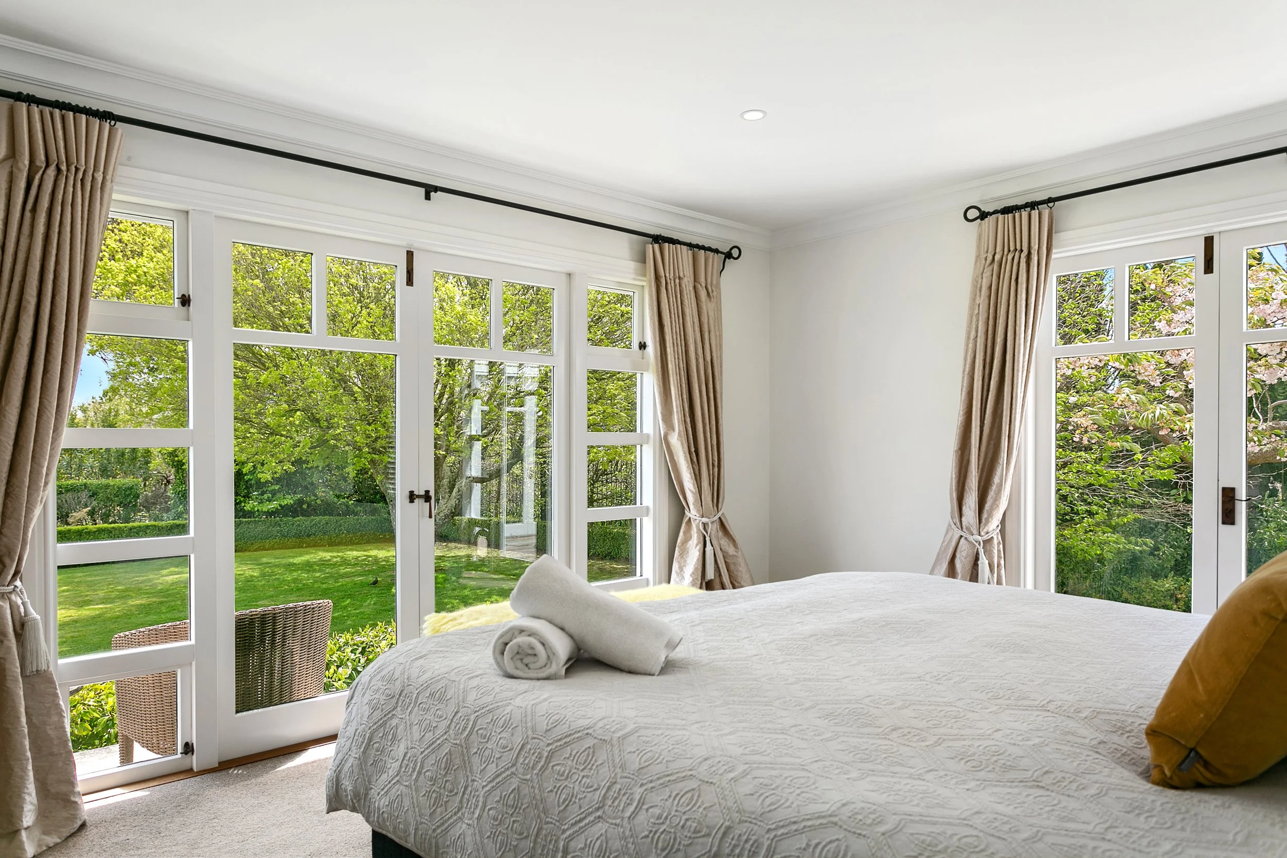 Bright bedroom with large windows revealing a lush green backyard, beige curtains, and a white bed with towels on top.