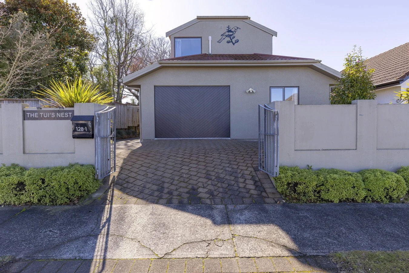Front of a modern gray house with a black garage door, surrounded by a white wall with a black mailbox and gate, and greenery on both sides.
