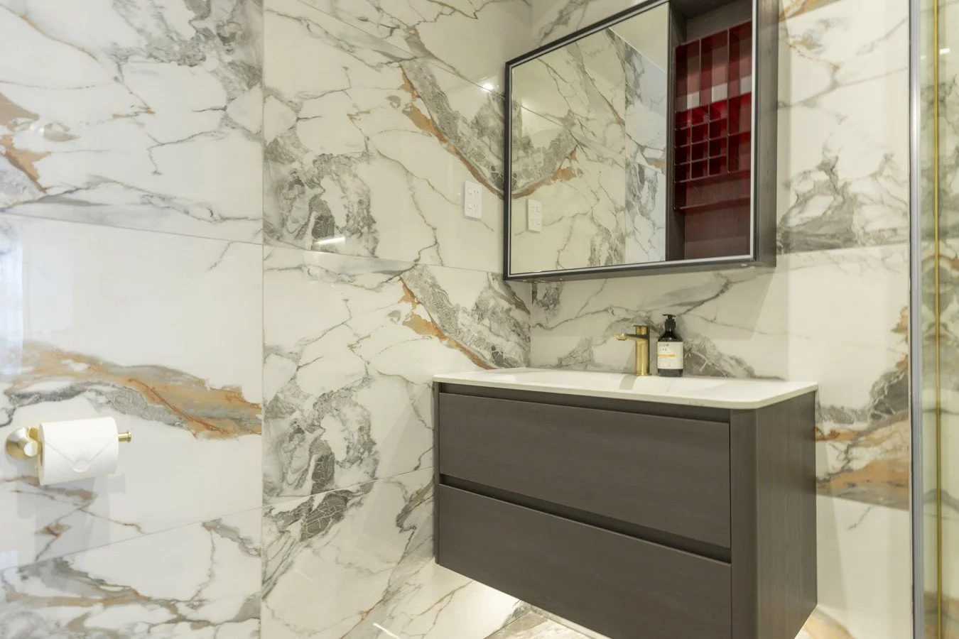 Bathroom with marble walls, a floating dark gray vanity with a white countertop, a gold faucet, a black soap dispenser, and a mirrored cabinet with wine rack inside.
