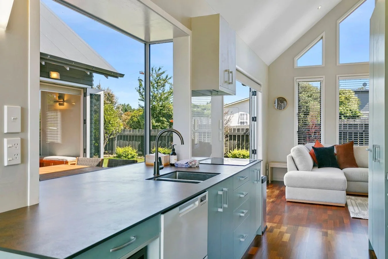 Bright kitchen with a large island, view through open glass doors to a backyard with trees, and a sitting area with a sofa and pillows inside.