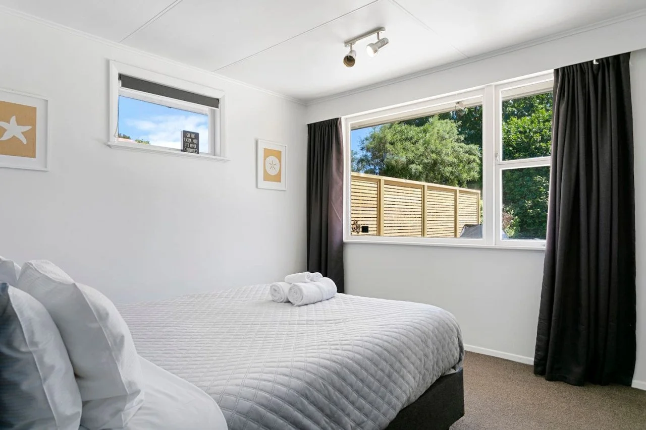 A bedroom with white walls, a bed with white bedding and pillows, and rolled towels on top. There are three framed pictures on the wall, large windows with black curtains, and trees outside.
