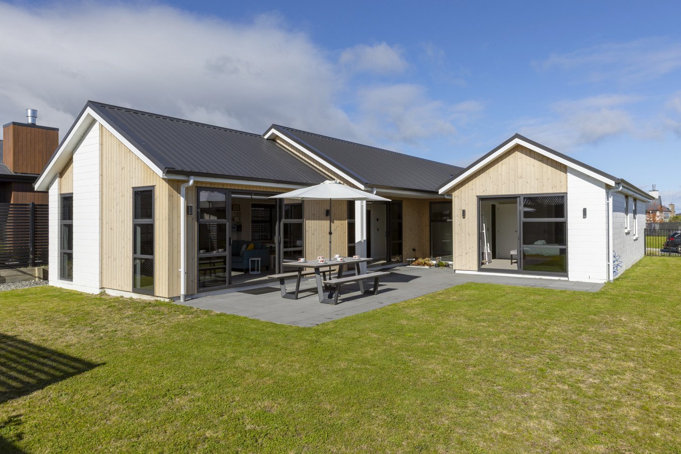Modern house with wooden and white exterior walls, black metal roofing, glass sliding doors, a patio with a picnic table and umbrella, and a grassy backyard under a partly cloudy sky.