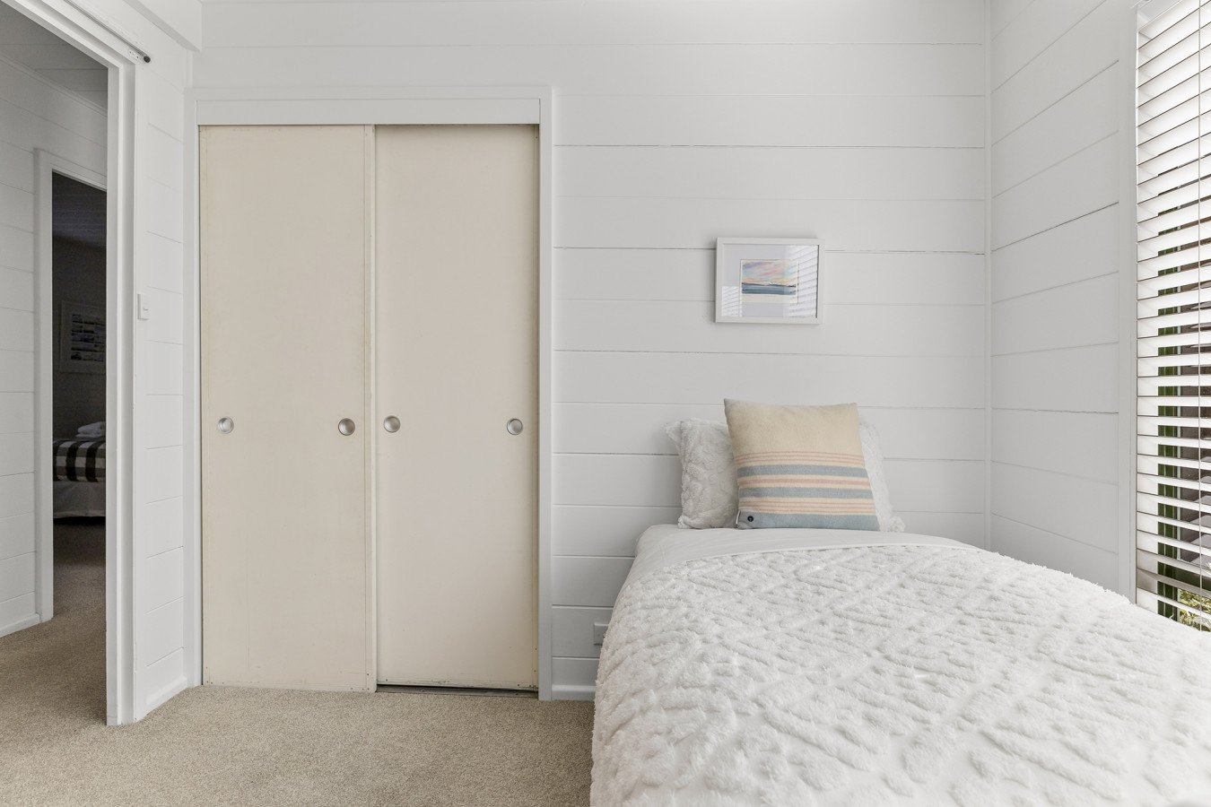 A bedroom with white shiplap walls, a closed closet door, a bed with white textured bedding, a striped pillow, a small framed artwork on the wall, and a window with blinds.