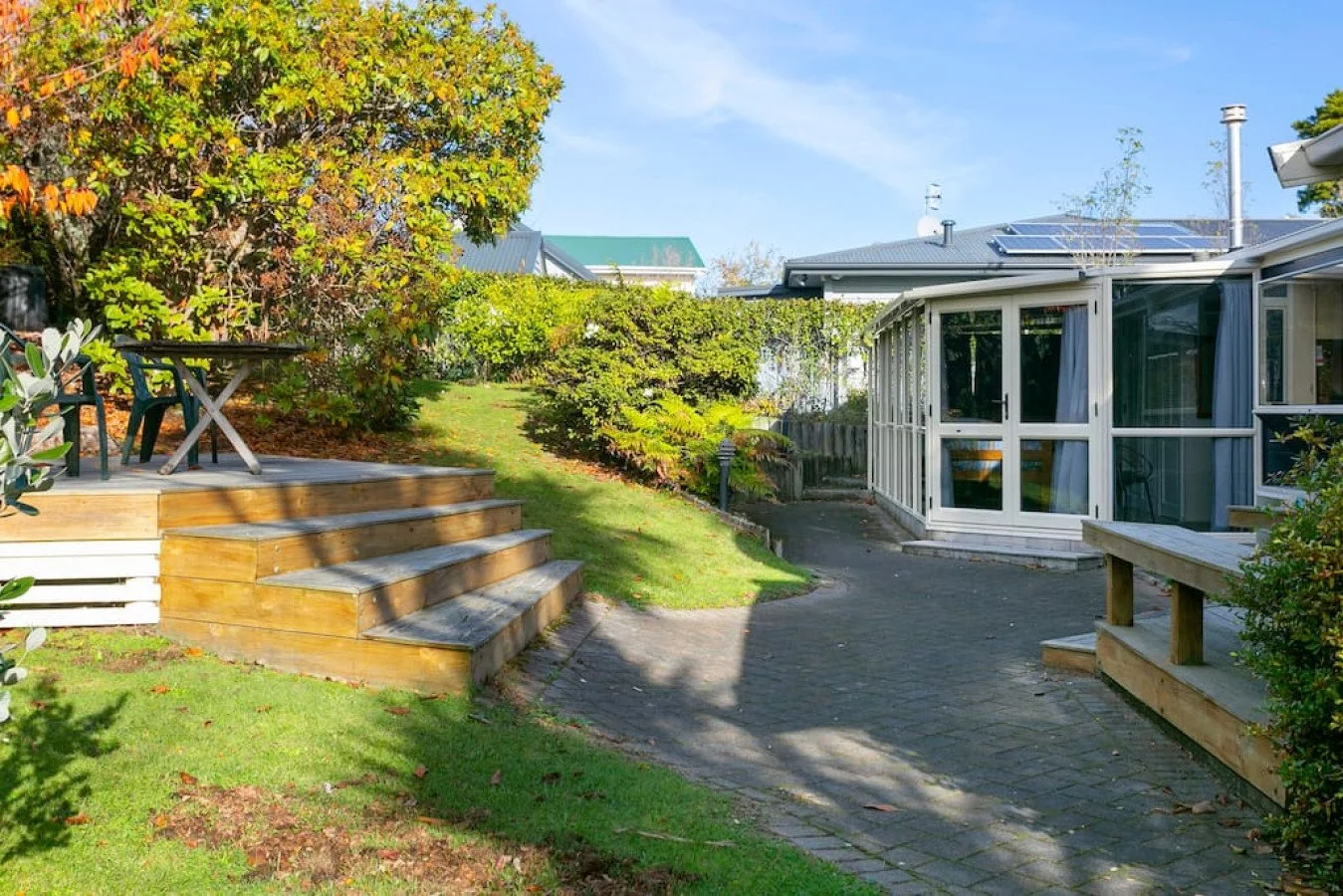 A backyard with a wooden deck with steps, tables and chairs, surrounded by lush green trees and bushes, and a house with a sunroom and solar panels on the roof under a clear blue sky.