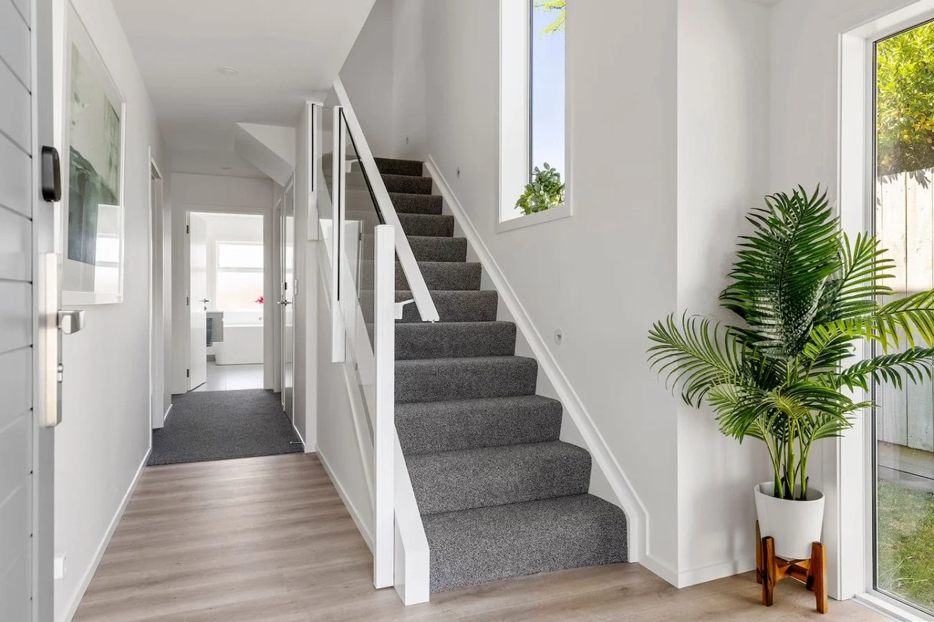Interior of a modern house with a staircase carpeted in gray, white walls, wooden flooring, and a large potted plant near a glass door.